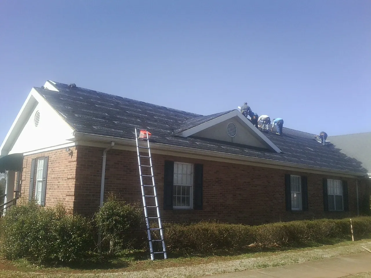 Crew tearing shingles off a brick ranch roof with a ladder set — Emergency Roof Repair work by our crew in Massena
