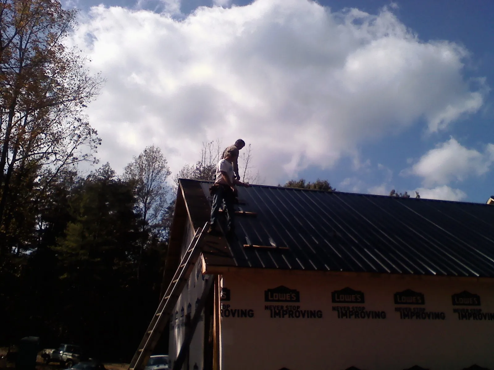 Roofer fastening dark metal panels high against a cloudy sky, from a Roof Inspection job in Massena